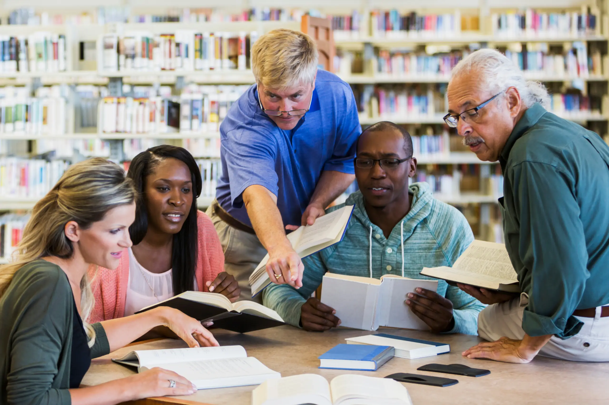 A diverse group of five adults studying together at a table in the library. They are having a discussion, with books open in their hands and on the table. The youngest is in his mid twenties, the oldest is late seventies.