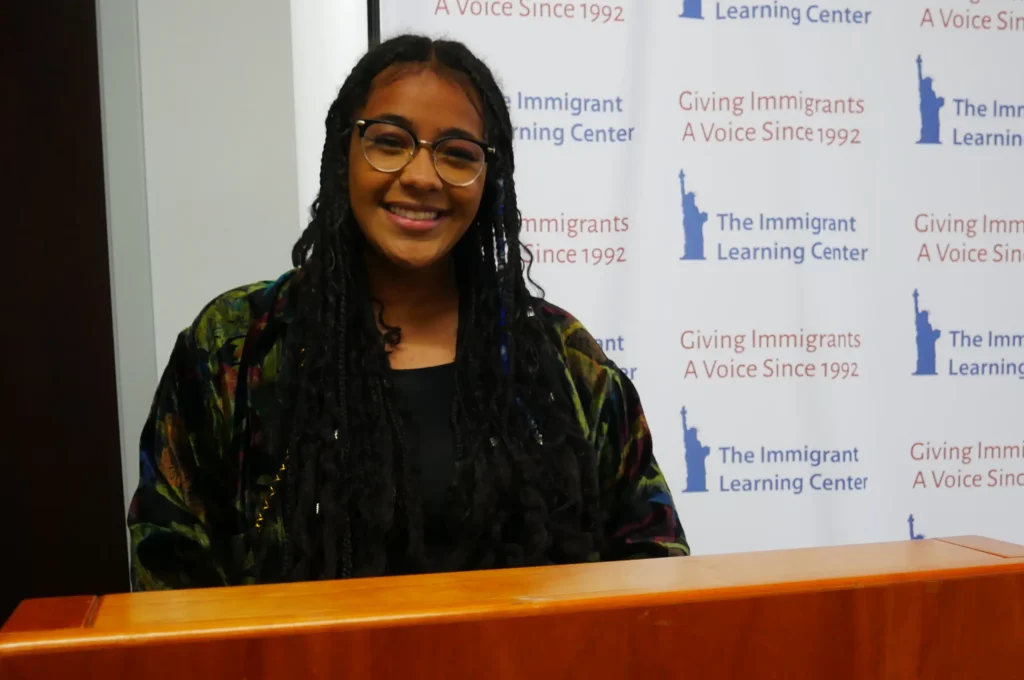 Woman smiling behind podium with The Immigrant Learning Center logo as a background