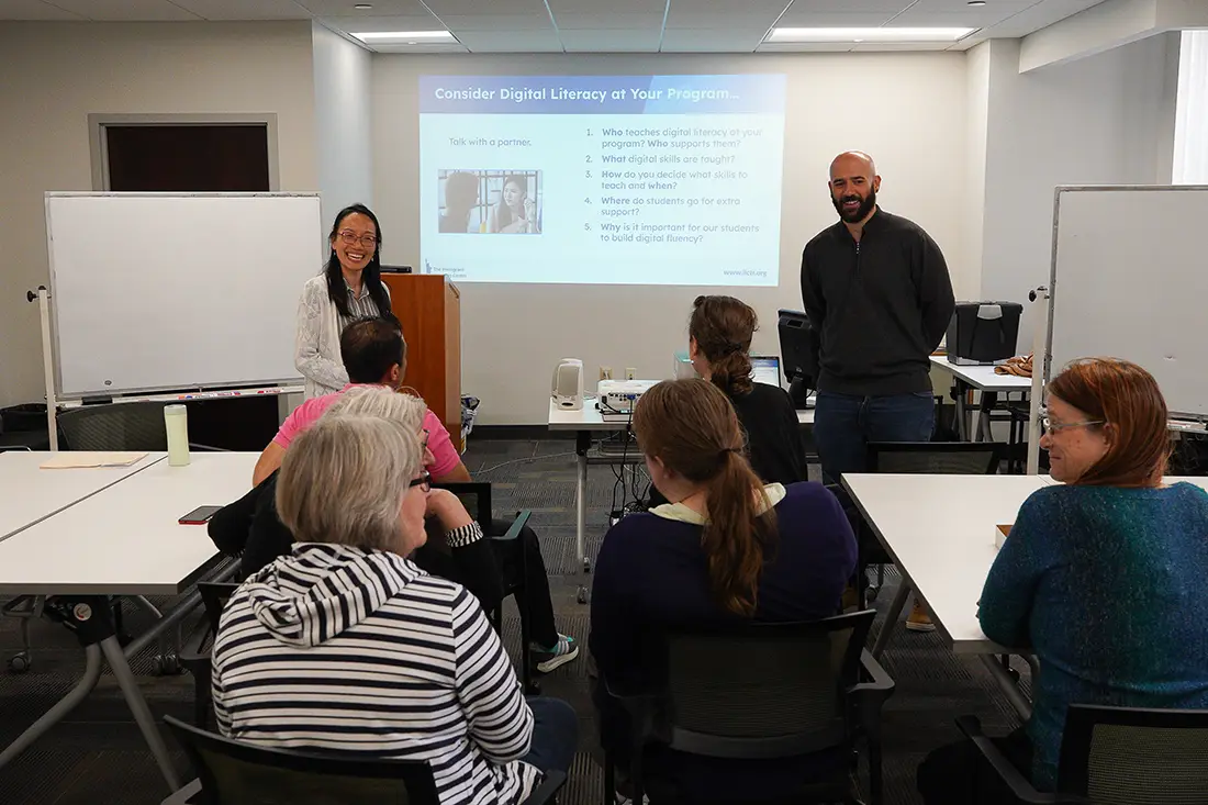 An ILC teacher and an advisor in a room with people at desks listening. A presentation is on the screen behind them.