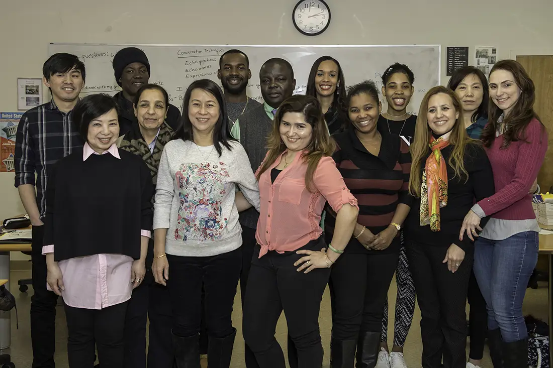 Fourteen students standing together in a classroom and smiling at the camera.