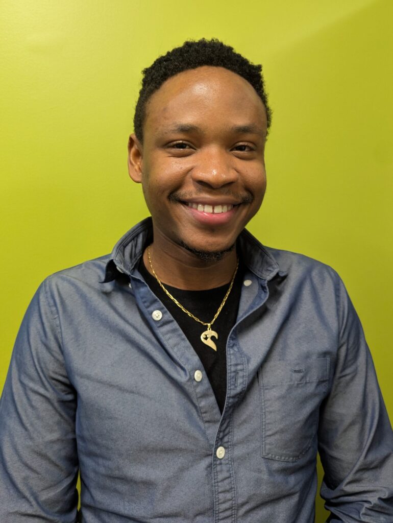 A man standing in front of a bright green background. He's wearing a dark gray button up shirt and he's smiling.