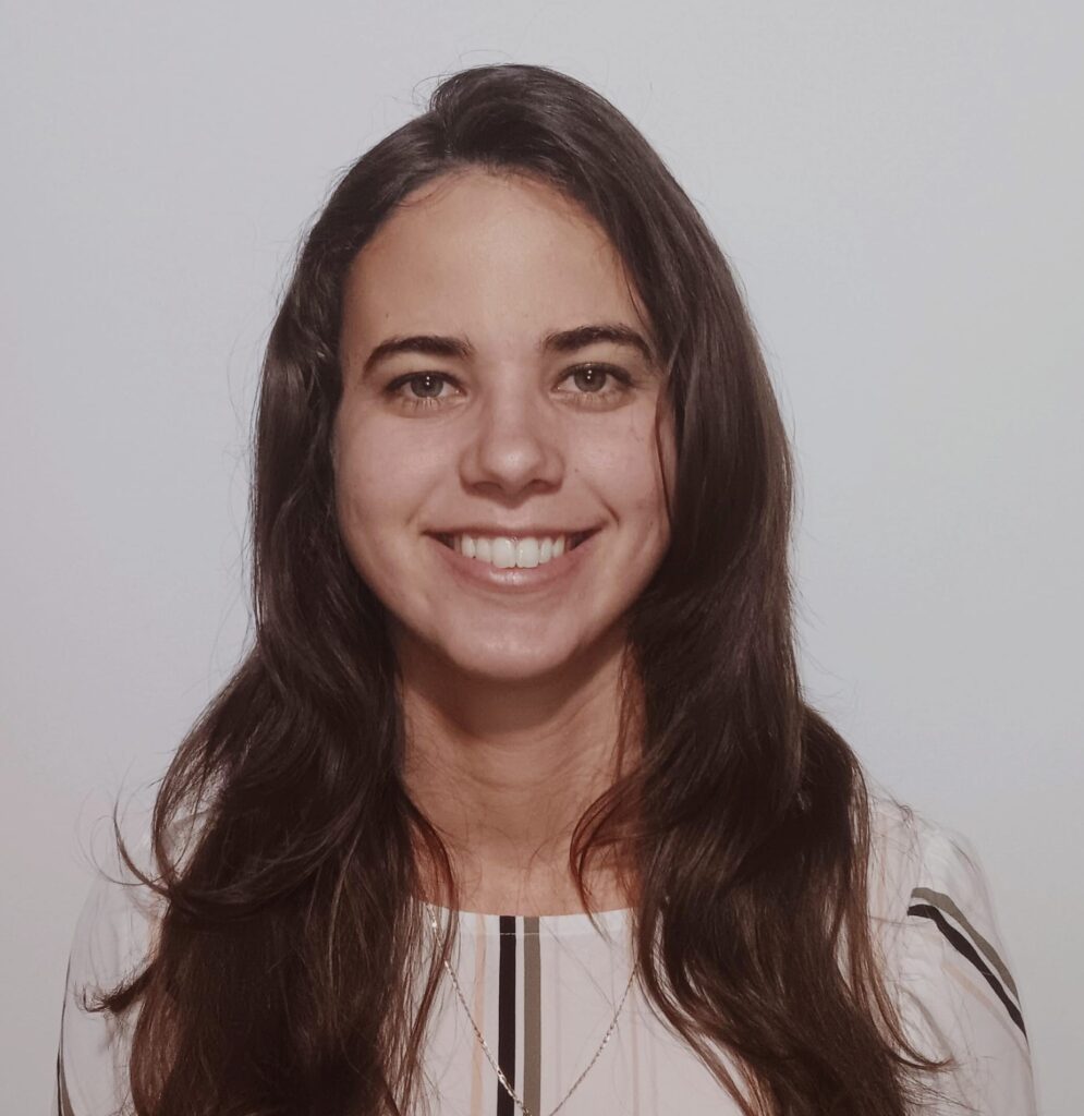 A woman with long brown hair standing in front of a light gray wall. She's wearing a white shirt and smiling.